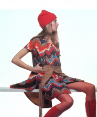 Model sitting on white slat wears bright red and blue aztec print sleeveless coat by Gianni Penati (1970)